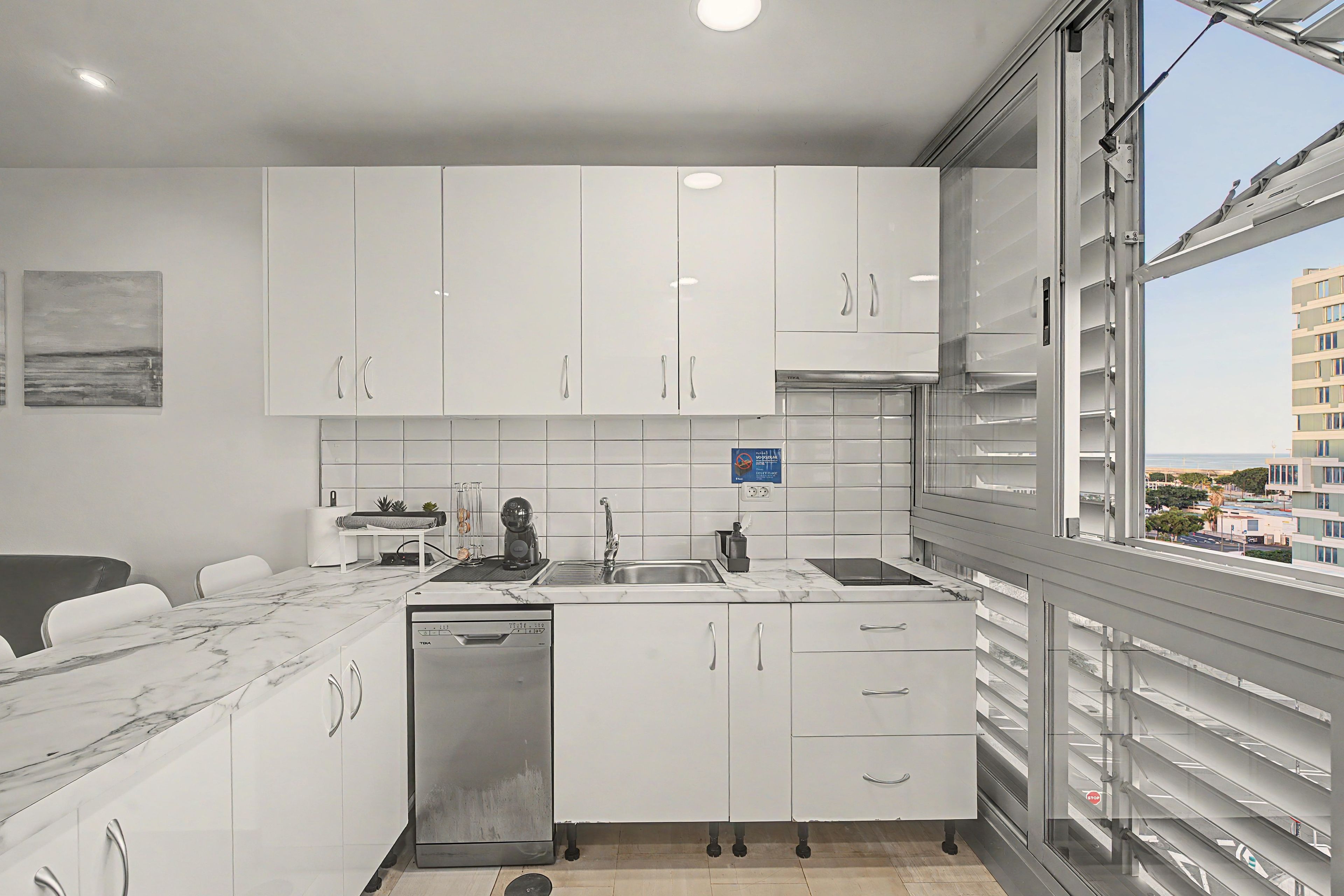 A modern kitchen with white cabinets, a dishwasher, and a view of the city through large windows.