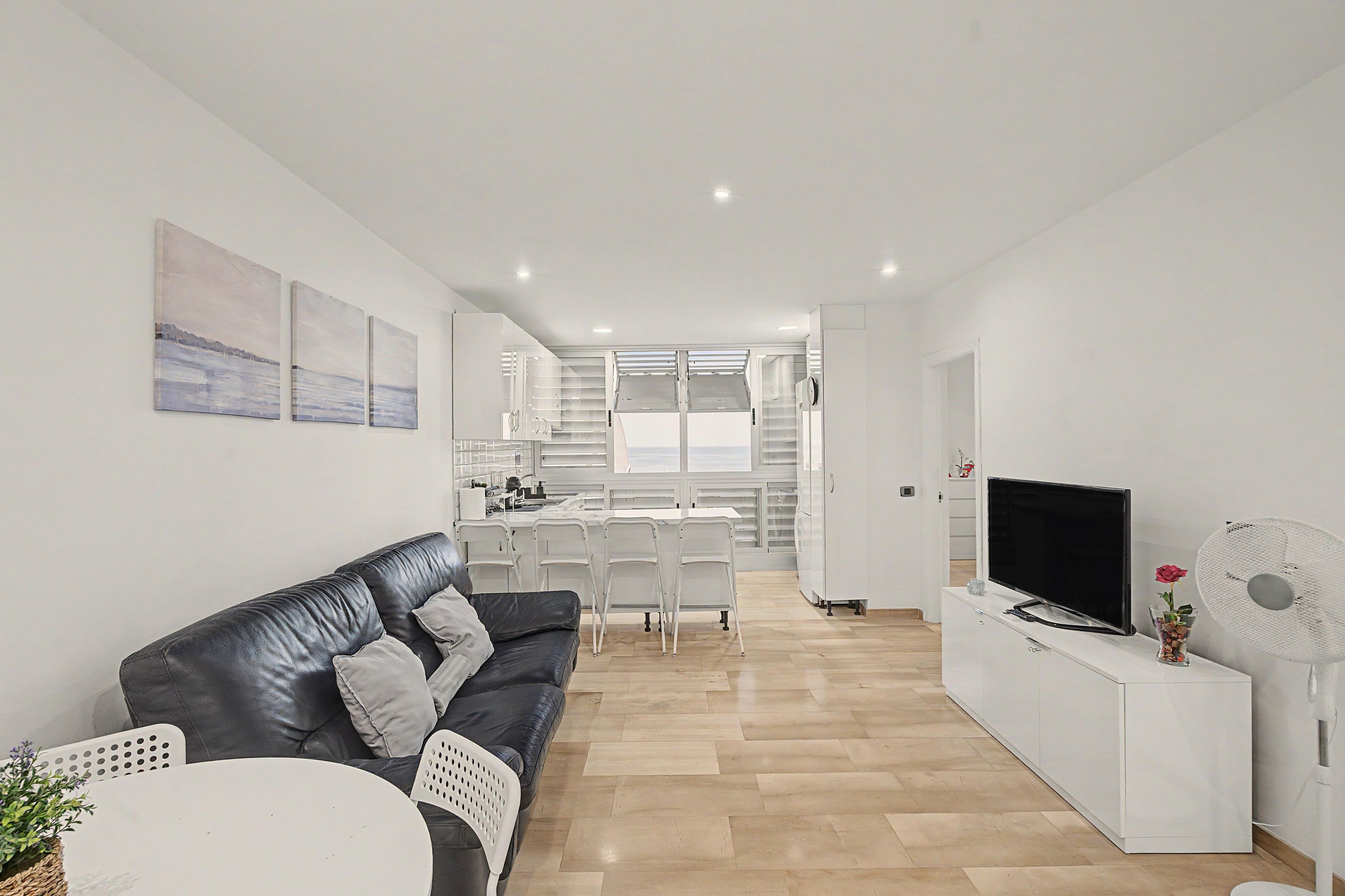 A modern living room with a black leather sofa, a dining area with white chairs, a kitchen with white cabinets, and a TV on a white stand.