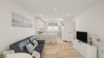 A modern living room with a black leather sofa, a dining area with white chairs, a kitchen with white cabinets, and a TV on a white stand.
