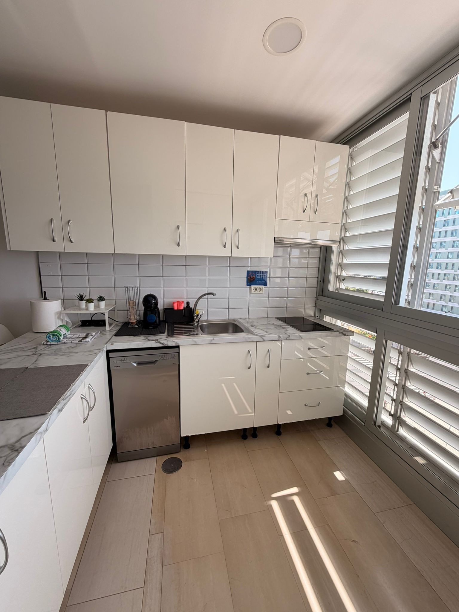 Una cocina moderna con armarios blancos, lavavajillas integrado y ventana con persianas.