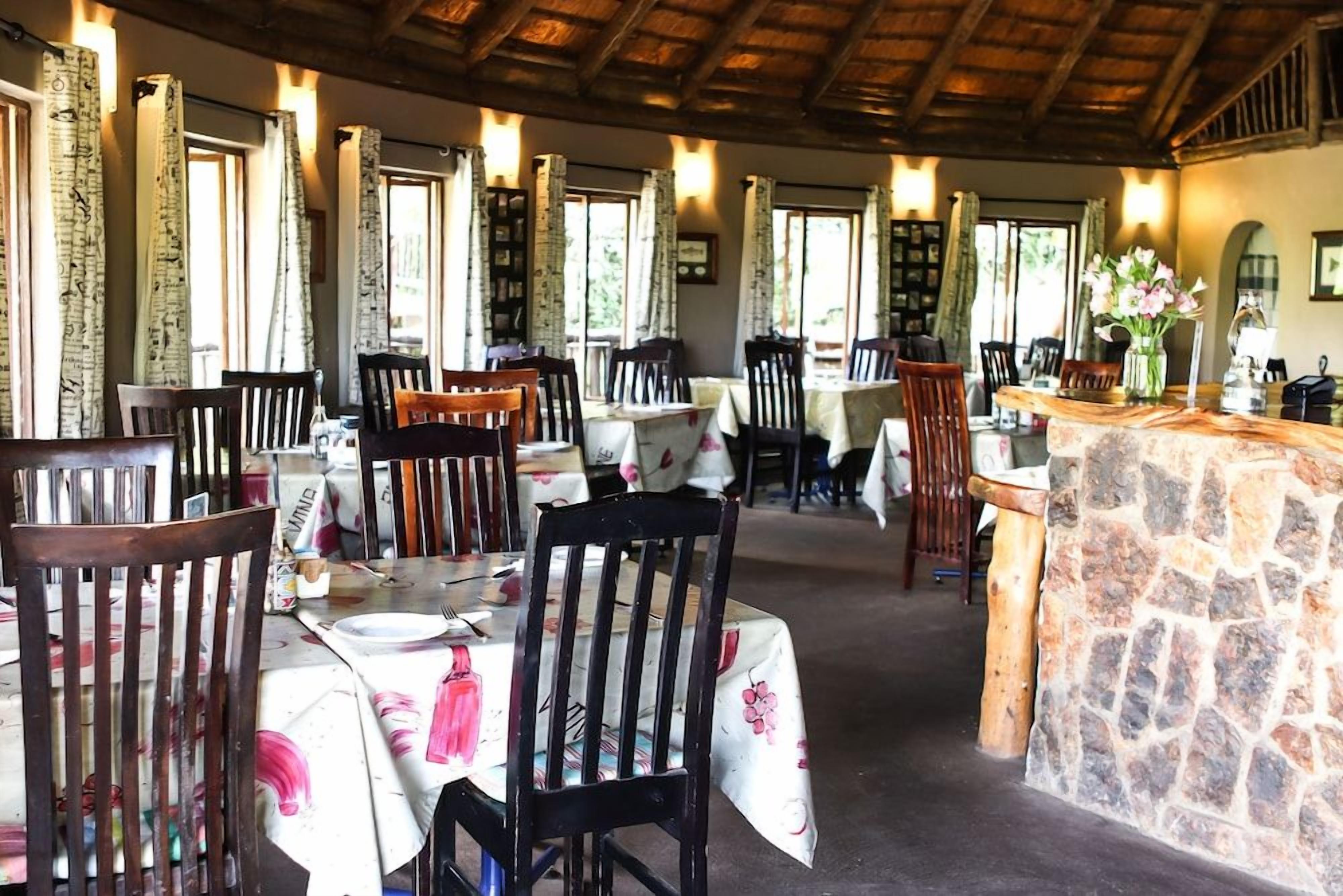 A restaurant with wooden chairs and tables, stone bar counter, and large windows with curtains.