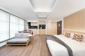 Una habitación de hotel moderna con un sofá, una mesita, una cama con almohadones estampados y una cocina americana al fondo.