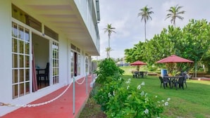 Un hôtel avec une entrée surmontée d’un tapis rouge, des tables à l’extérieur avec des parasols rouges et des palmiers en arrière-plan.