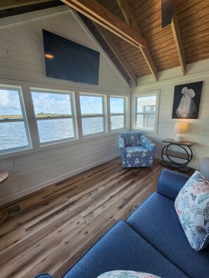 A room with a wooden floor, a blue sofa, a patterned armchair, a table with a lamp, and a painting on the wall.