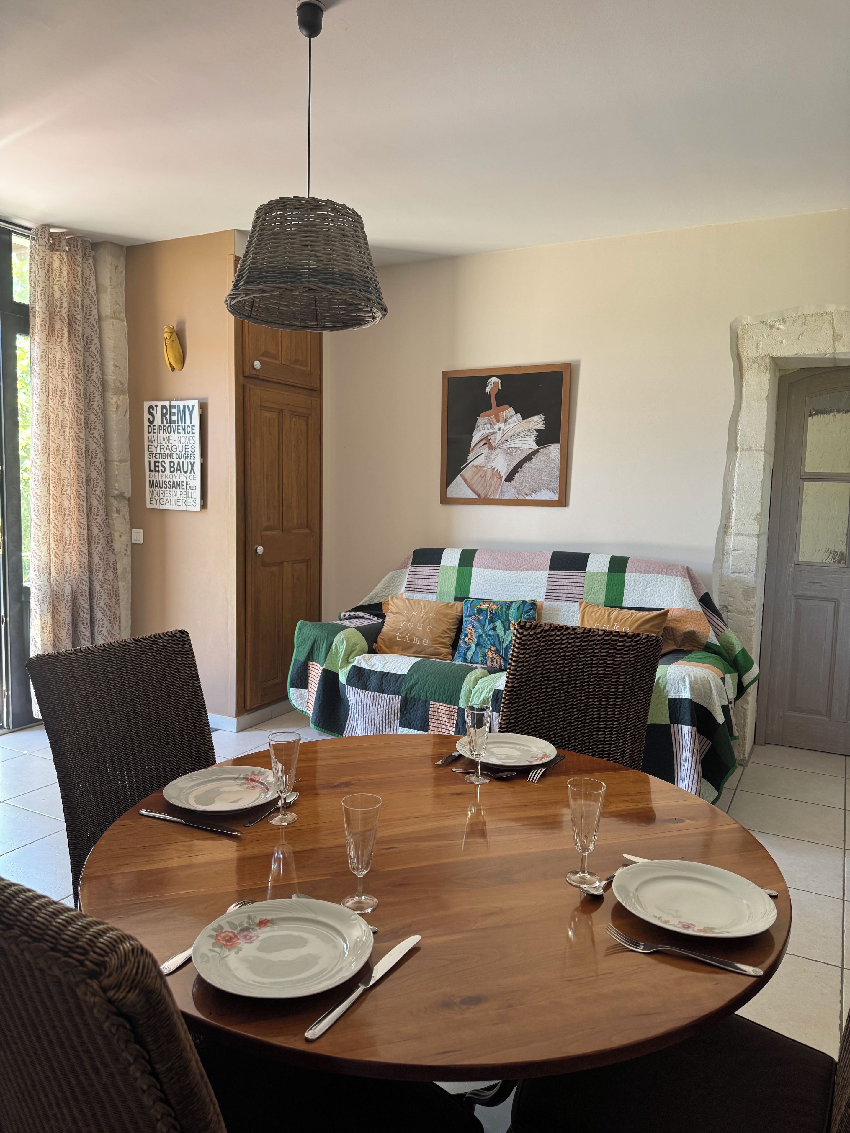 Un coin repas avec une table ronde en bois, dressée pour quatre personnes, une chaise en osier et un canapé recouvert d’une courtepointe à motifs.