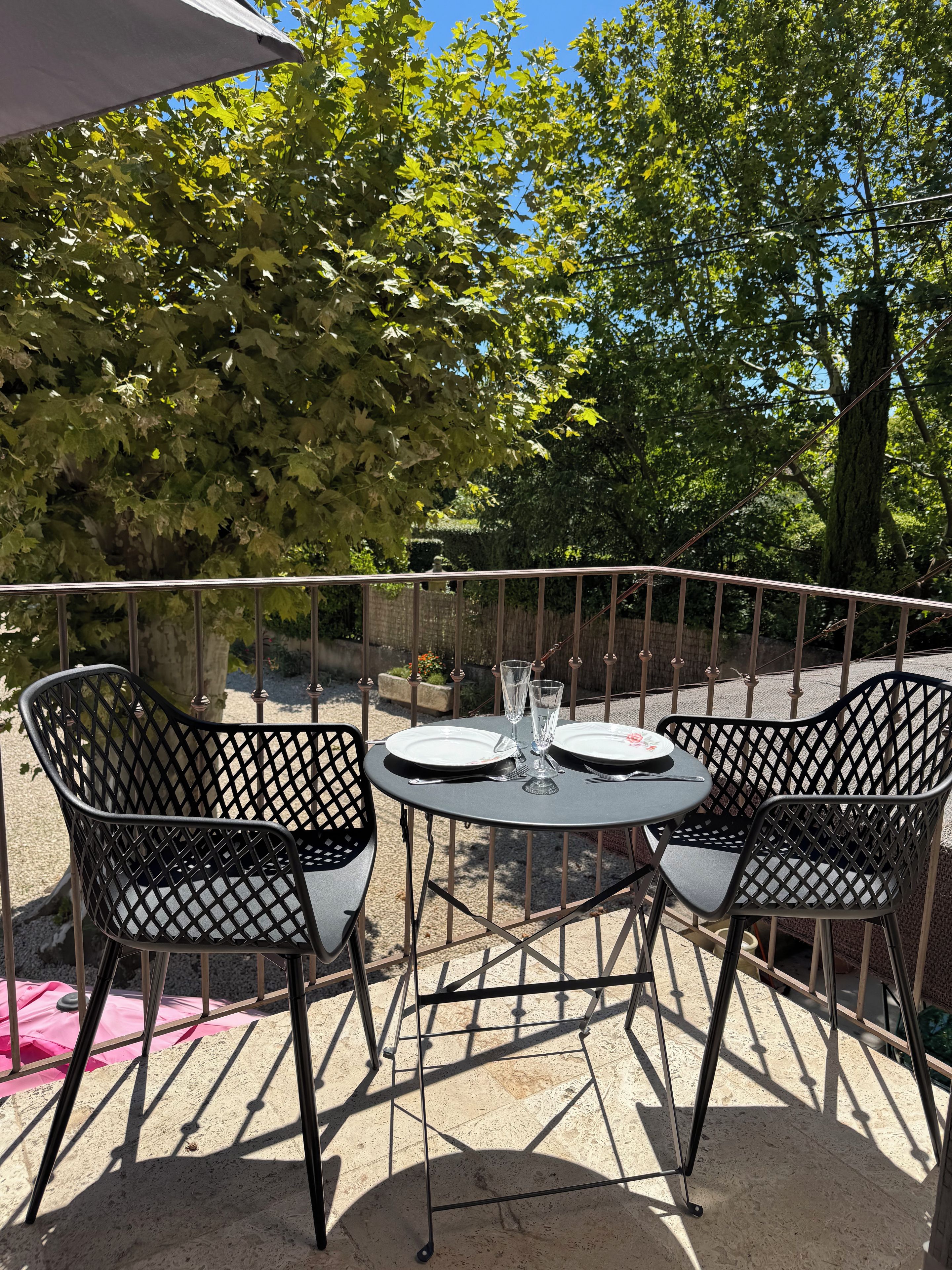 A sunlit outdoor dining area with two metal chairs and a table, surrounded by a metal railing and trees.