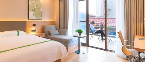 A hotel room with a bed, a sofa, a small table, and a chair. There is a large window with a view of the outside.