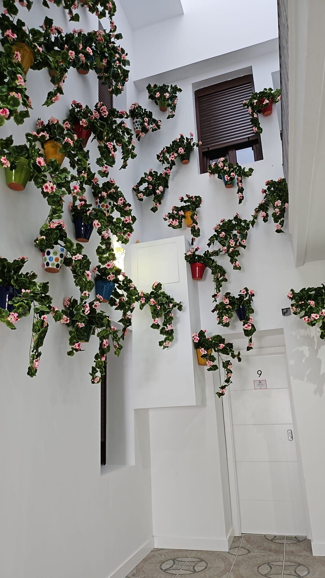 Un pasillo con varias plantas en macetas colgando del techo, una puerta con número y una ventana con persianas.