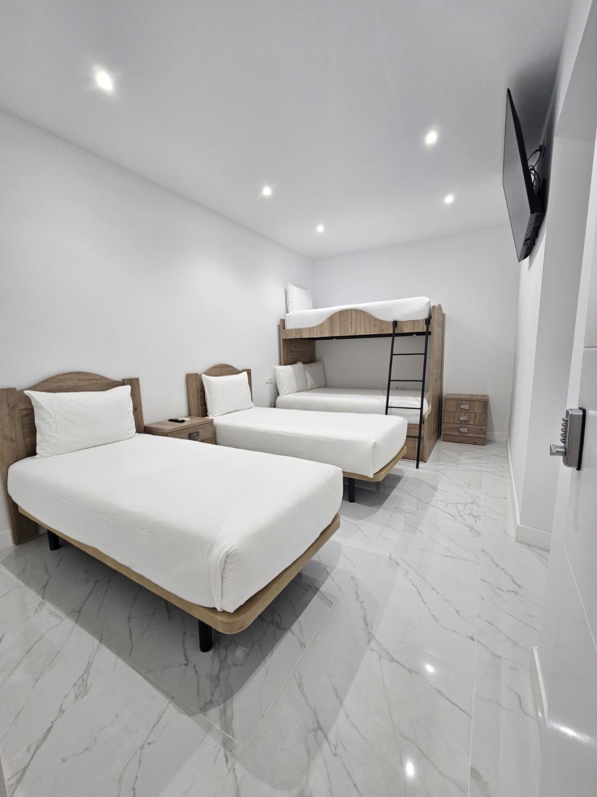 Un dormitorio moderno con dos camas individuales, una litera, un televisor montado en la pared y suelo de mármol.