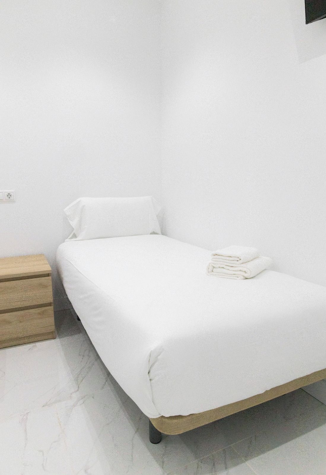 A single-bed room with a wooden nightstand, a folded white towel, and a wall socket.