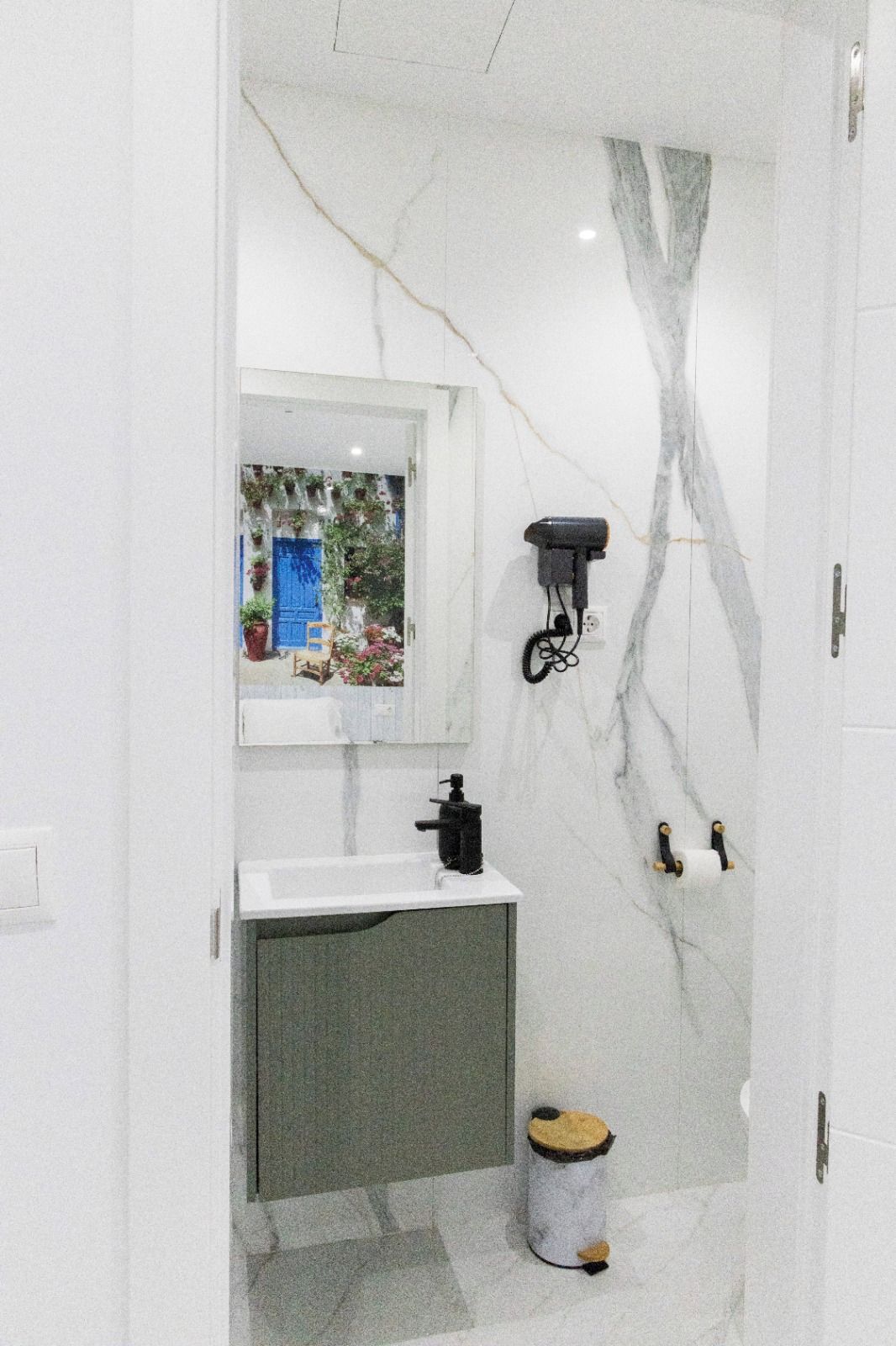 A bathroom with a white marble wall, a sink with a mirror, a hairdryer mounted on the wall, and a toilet with a bidet attachment.