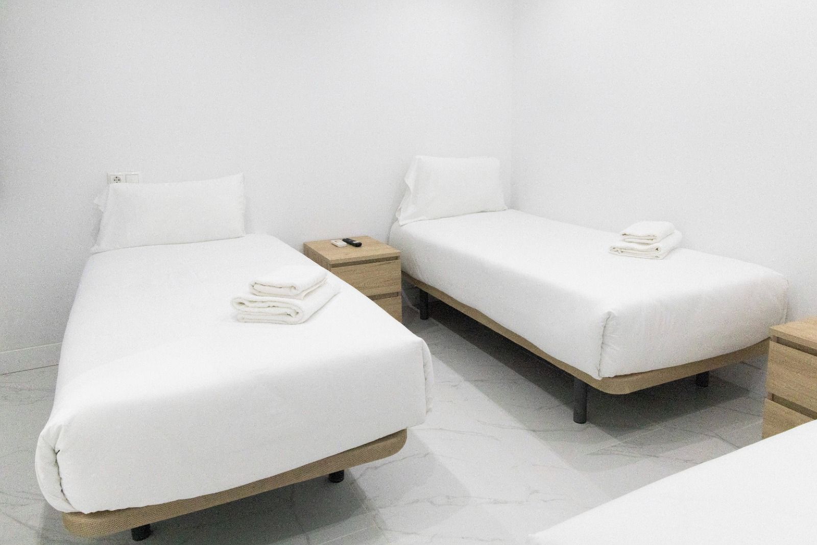 A hotel room with two single beds, each with a wooden frame and white bedding. There is a small bedside table with a lamp and a folded towel on each bed.
