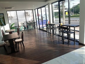 A spacious indoor area with multiple tables and chairs, large windows, and a view of the street outside. - NK Residence Sakon Nakhon (Sakon Nakhon)