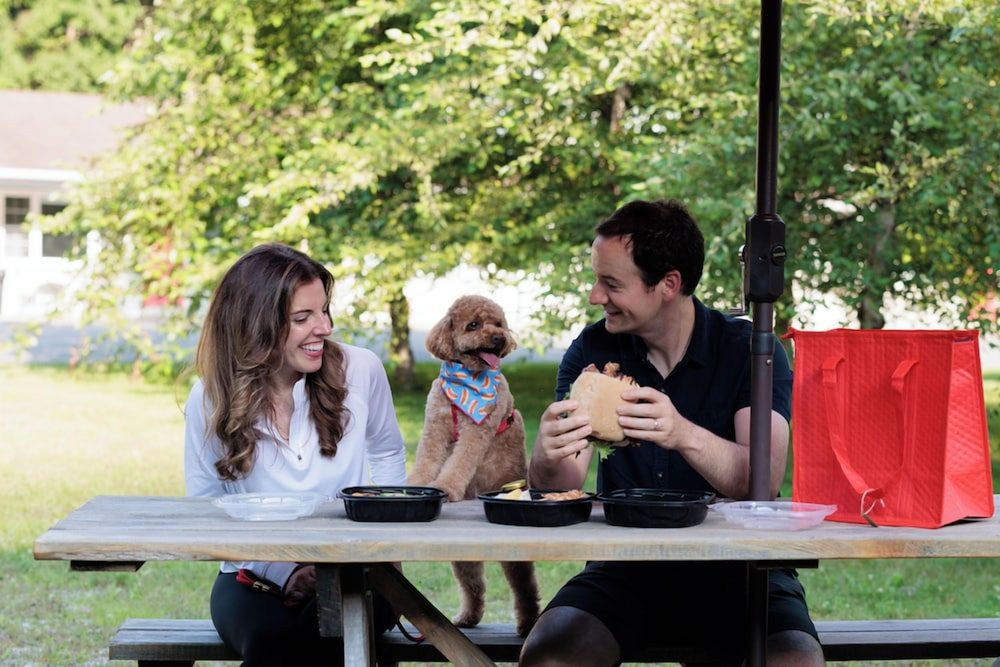 A picnic table with food containers, a red bag, and a person eating.