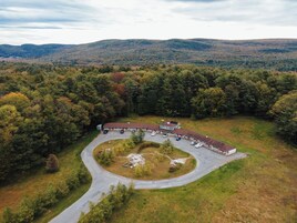 A large building with a parking lot and a driveway, surrounded by dense forest and hills.