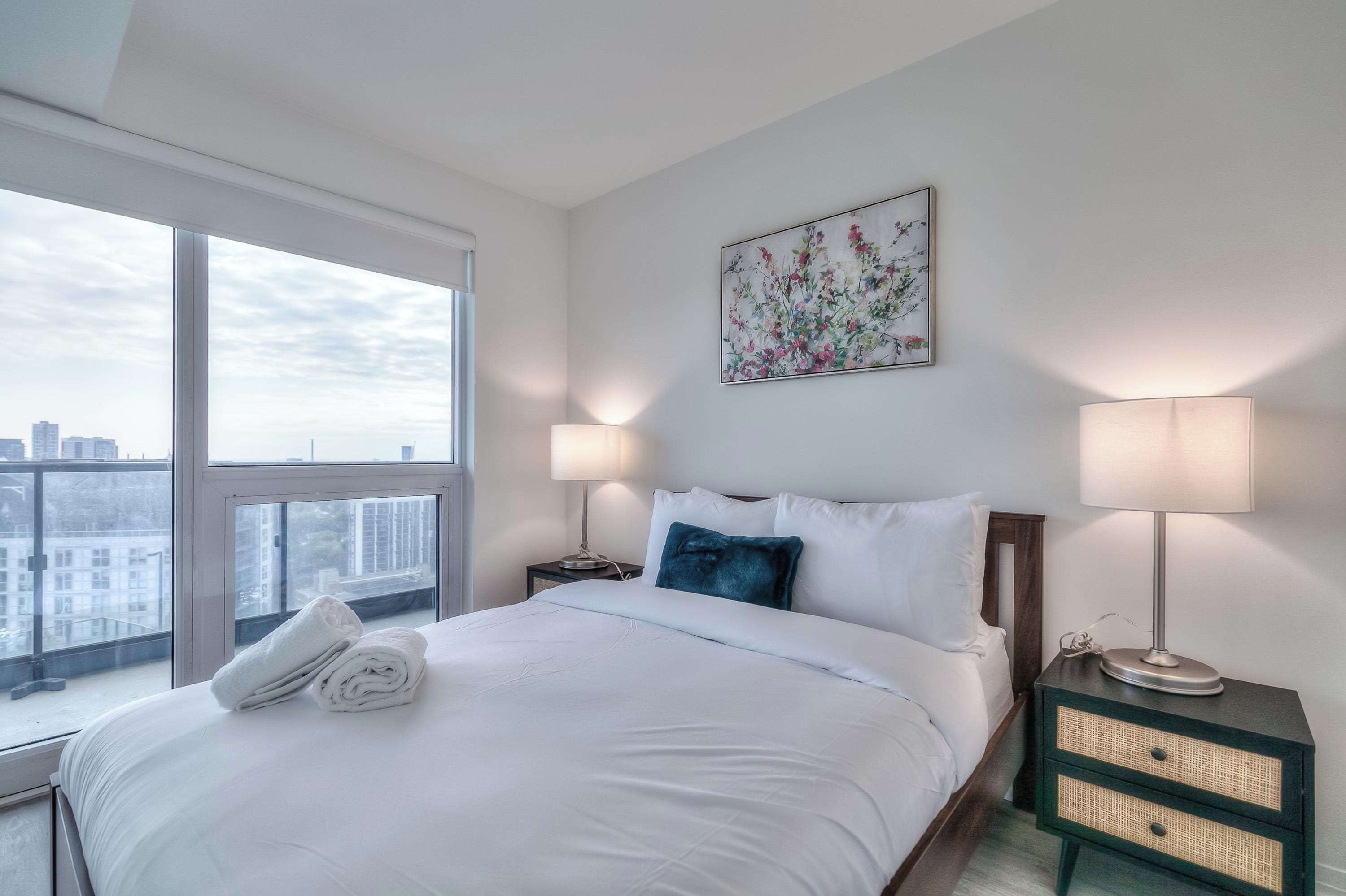 Habitación de hotel con un ventanal grande, cama con ropa blanca, almohada azul, dos lámparas de noche y vista a la ciudad.