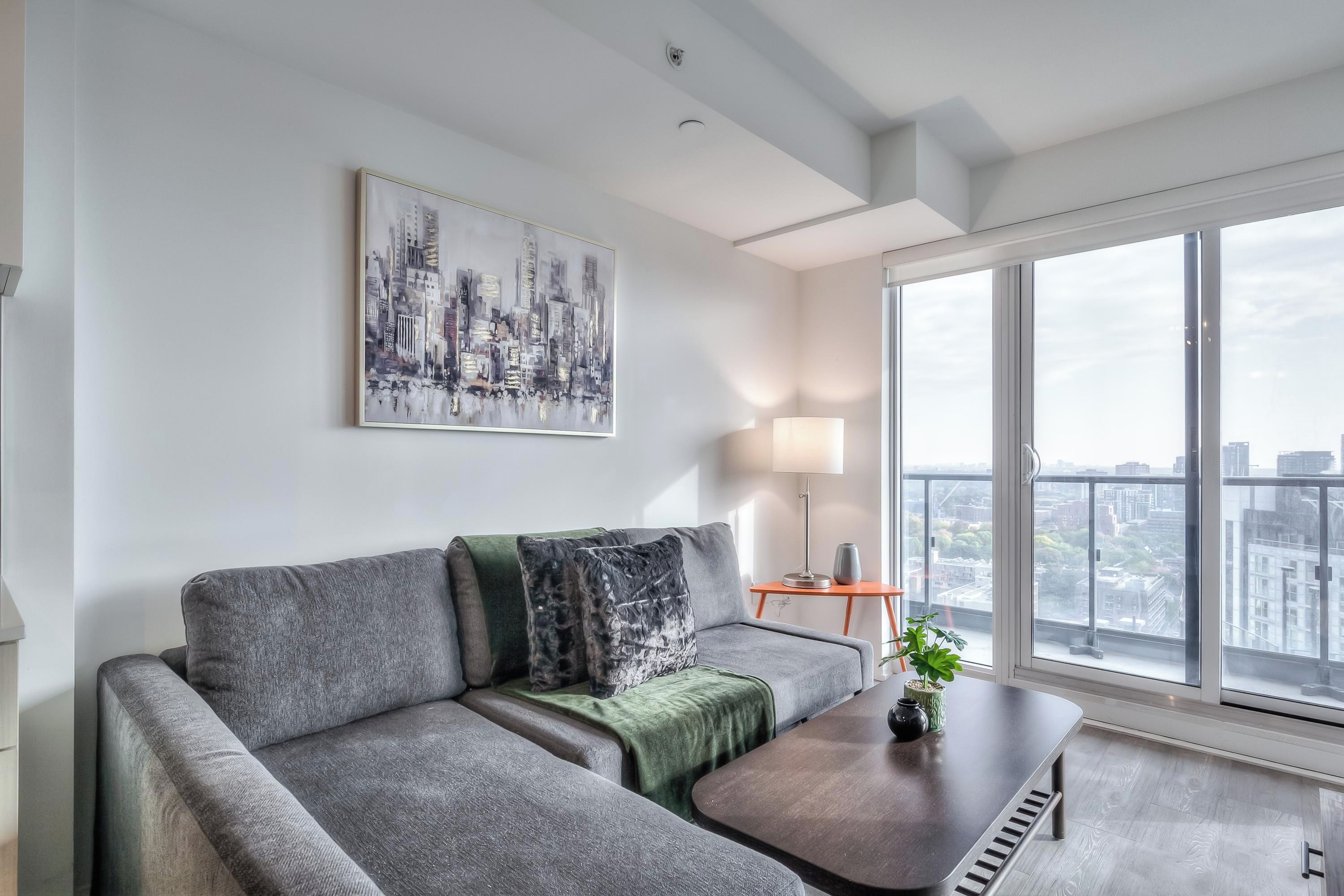 Una sala de estar moderna con un sofá gris, una manta verde, una mesa de centro y vistas a la ciudad a través de amplias ventanas.