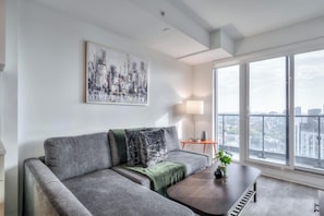 Un salon moderne avec un canapé gris, un plaid vert, une table basse et une vue sur la ville à travers de grandes fenêtres.