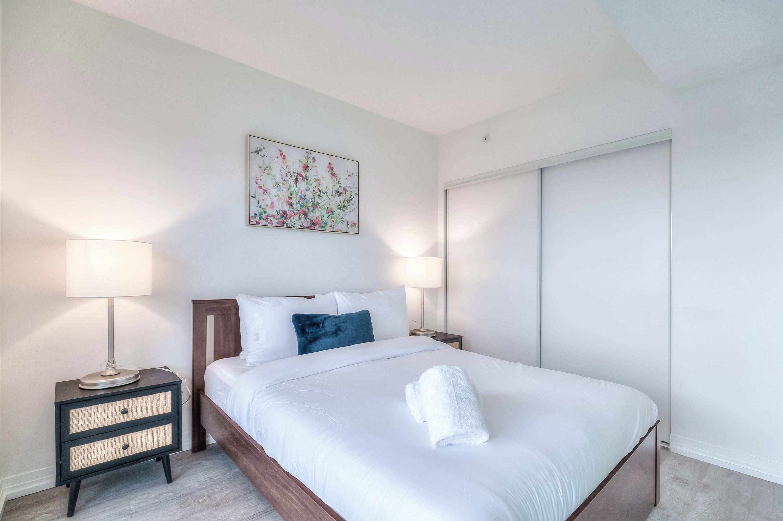 Una cama bien hecha con sábanas blancas, un cojín azul y una toalla blanca sobre la cama. Hay una mesita de noche con una lámpara y un cajón pequeño. Un cuadro cuelga en la pared sobre la cama.