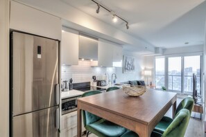 Une cuisine moderne avec un réfrigérateur en acier inoxydable, une table à manger en bois et des chaises à revêtement vert, et une vue sur la ville à travers de grandes fenêtres.