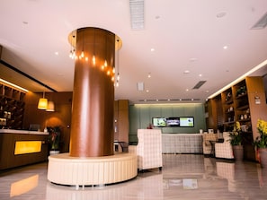 Un hall d’hôtel moderne doté d’une colonne cylindrique centrale, d’un espace salon, d’un comptoir d’accueil et d’écrans de télévision.