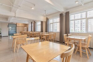 A spacious dining area with multiple wooden tables and chairs, large windows with curtains, and a modern kitchen in the background. - Borrman Wanda Plaza Houjie Dongguan (Dongguan)
