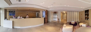 Un espace d’accueil d’hôtel comprenant un comptoir incurvé, un bureau d’accueil, un écran numérique et des coins salon avec des tables rondes.