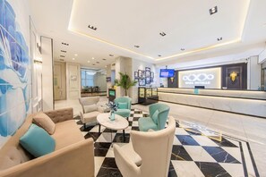 A hotel lobby with a checkered floor, seating area with sofas and armchairs, a reception desk, and a large wall with framed artwork. - Lavande Hotel (Tianjin)