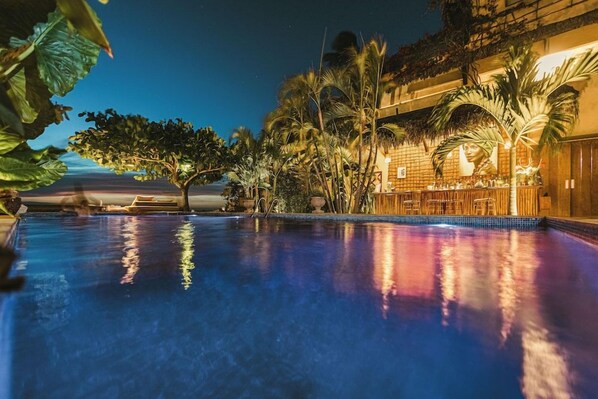 An indoor swimming pool with a tiled edge, surrounded by palm trees and a building with a brick facade. - Casa de Areia Jeri (Jijoca de Jericoacoara)