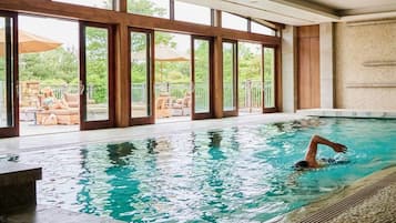 An indoor swimming pool with clear glass walls, a person swimming, and a view of outdoor seating.
