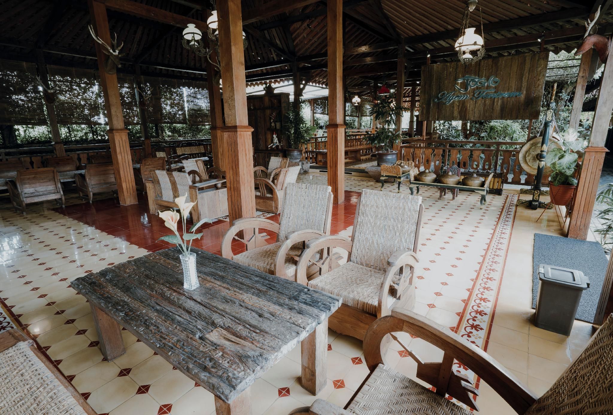 A covered outdoor seating area with wooden tables and chairs, potted plants, and a wooden sign.
