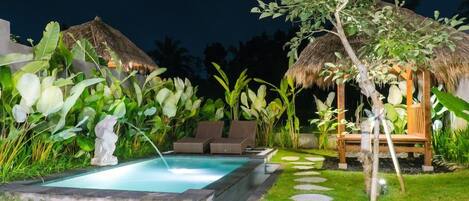 A pool area with lounge chairs, a thatched-roof structure, and tropical plants.