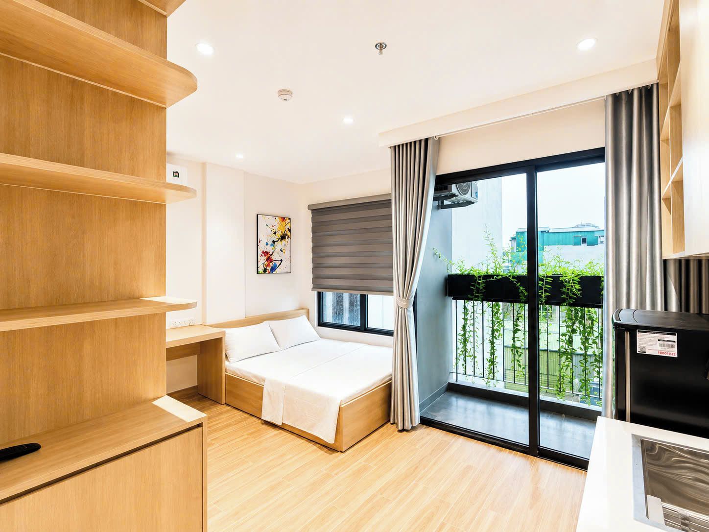 Habitación de hotel moderna con una cama, estanterías de madera, un cuadro, un balcón con vistas y un cubo de basura negro.