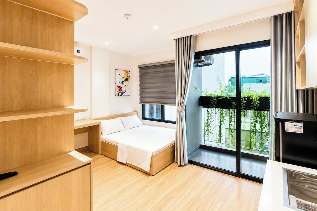 Quarto de hotel moderno com cama, estantes de madeira, um quadro, varanda com vista e um cesto de lixo preto.