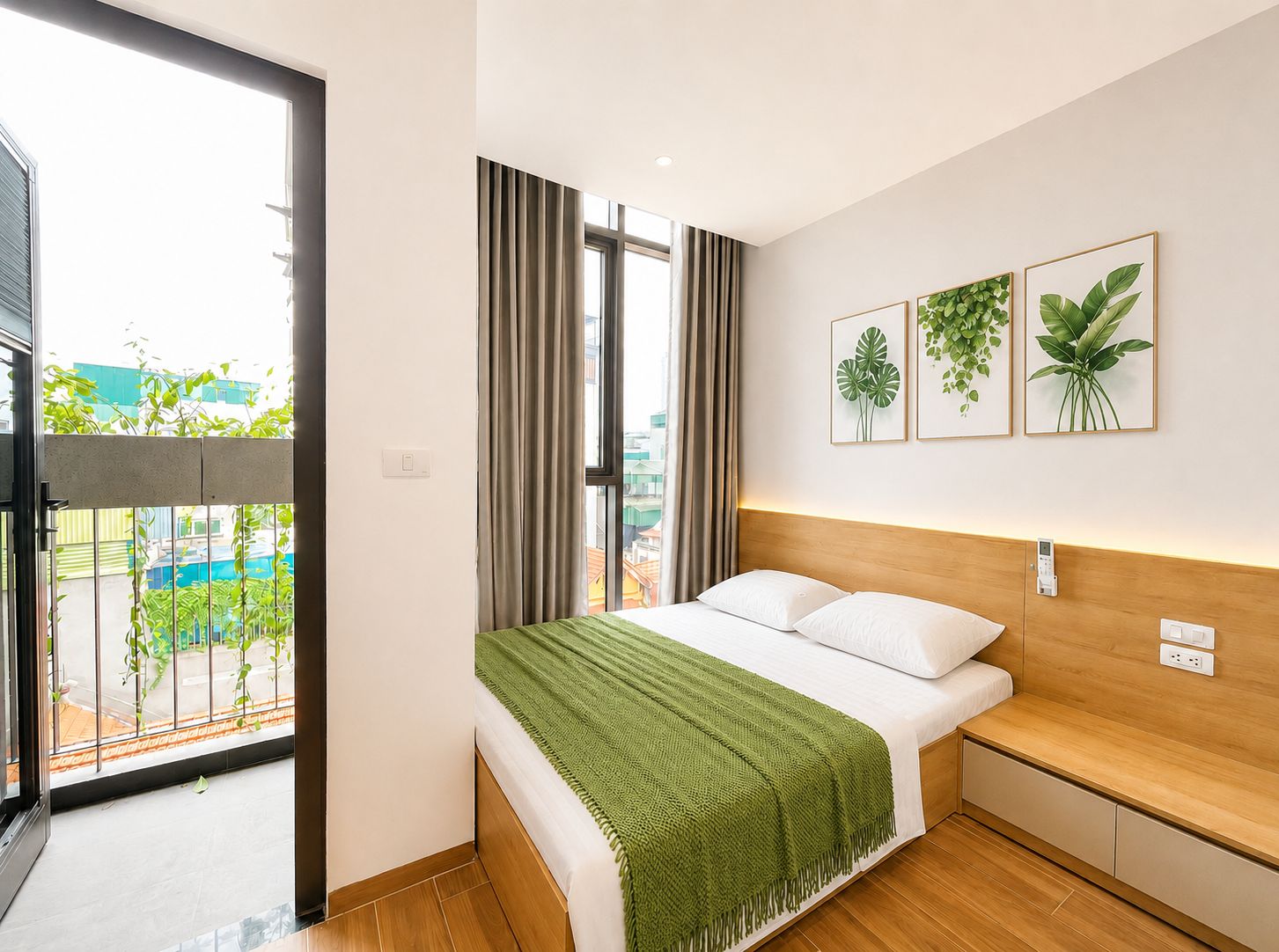 Une chambre d’hôtel moderne avec une grande fenêtre, un lit agrémenté d’un plaid vert et trois reproductions encadrées de motifs végétaux accrochées au mur.