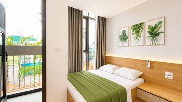 Une chambre d’hôtel moderne avec une grande fenêtre, un lit agrémenté d’un plaid vert et trois reproductions encadrées de motifs végétaux accrochées au mur.