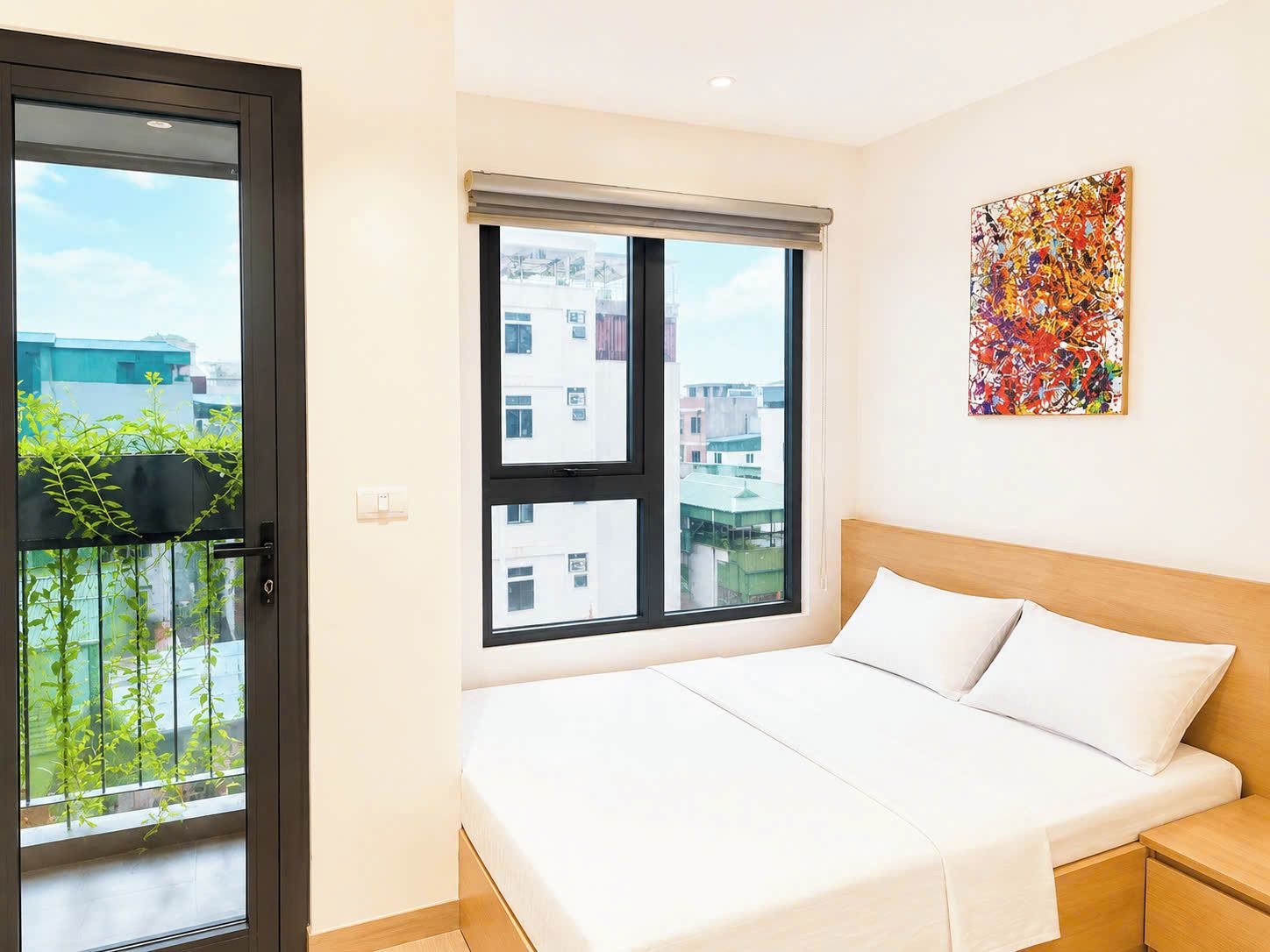 Quarto de hotel com uma cama, janela com vista para edifícios, um quadro na parede e uma varanda com plantas.
