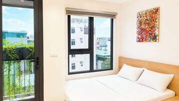 Quarto de hotel com uma cama, janela com vista para edifícios, um quadro na parede e uma varanda com plantas.