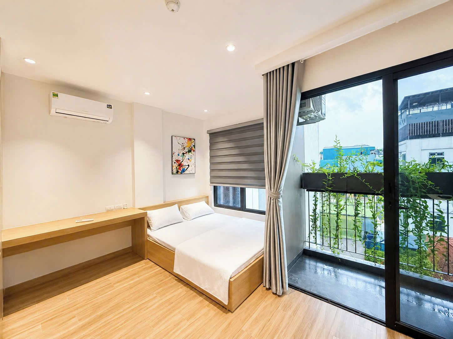 Une chambre d’hôtel moderne, équipée d’un lit, d’une table de chevet et d’une grande fenêtre offrant une vue sur les bâtiments et la verdure.
