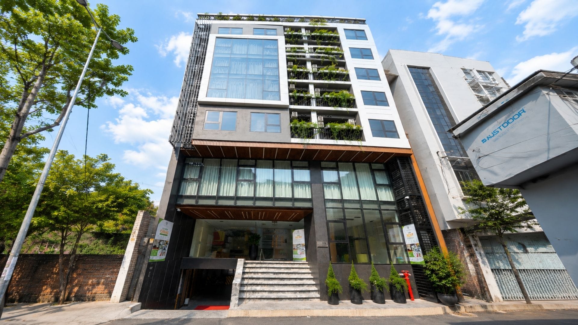 Un immeuble à plusieurs étages, doté d’un style architectural moderne, caractérisé par un mélange d’éléments en verre et en métal, une entrée bien visible avec des marches et un petit espace jardin aménagé aux étages supérieurs.