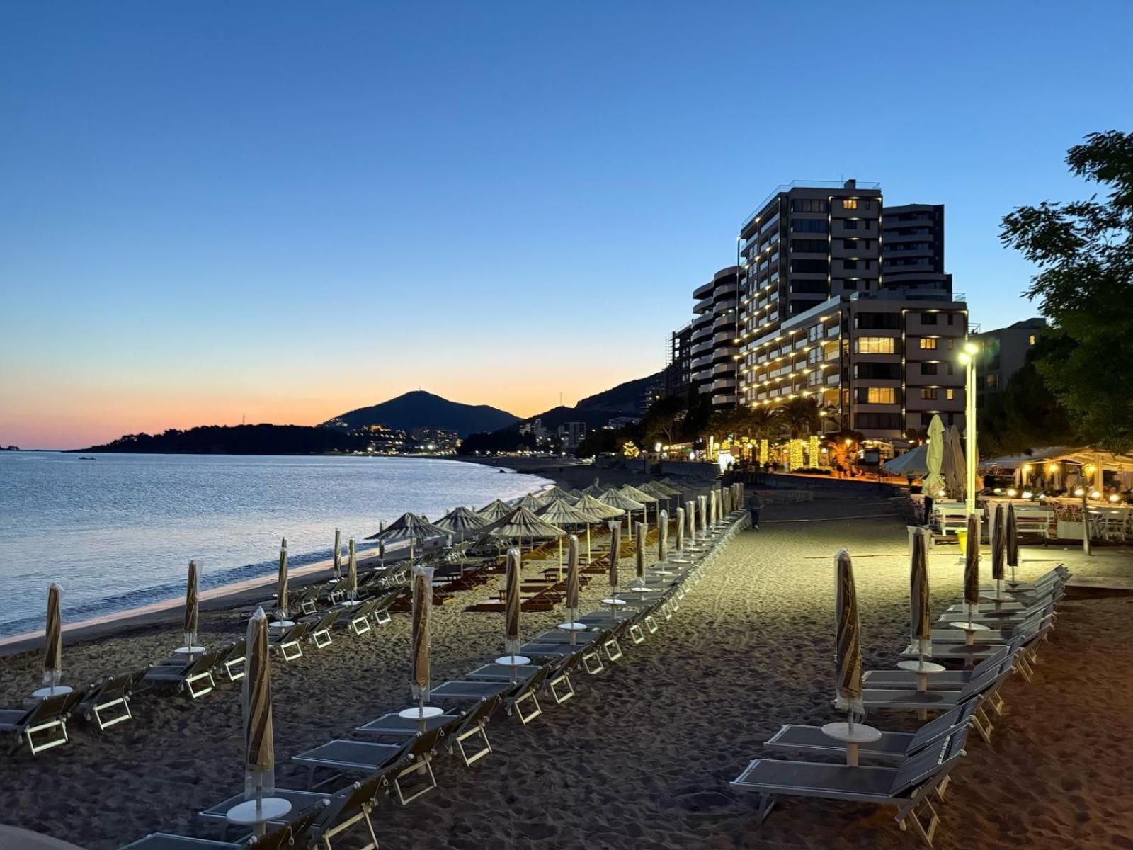 Un hotel frente a la playa con una fila de tumbonas y sombrillas, una playa y una montaña al fondo.