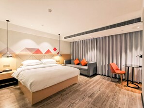 Quarto de hotel com uma cama, um sofá, uma cadeira, uma mesinha e uma cortina.
