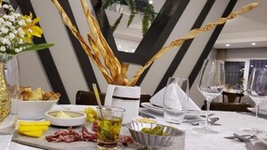 Una mesa de comedor dispuesta con una copa de cristal transparente, un cuenco de aceitunas, un vaso de líquido amarillo y un florero con flores blancas.
