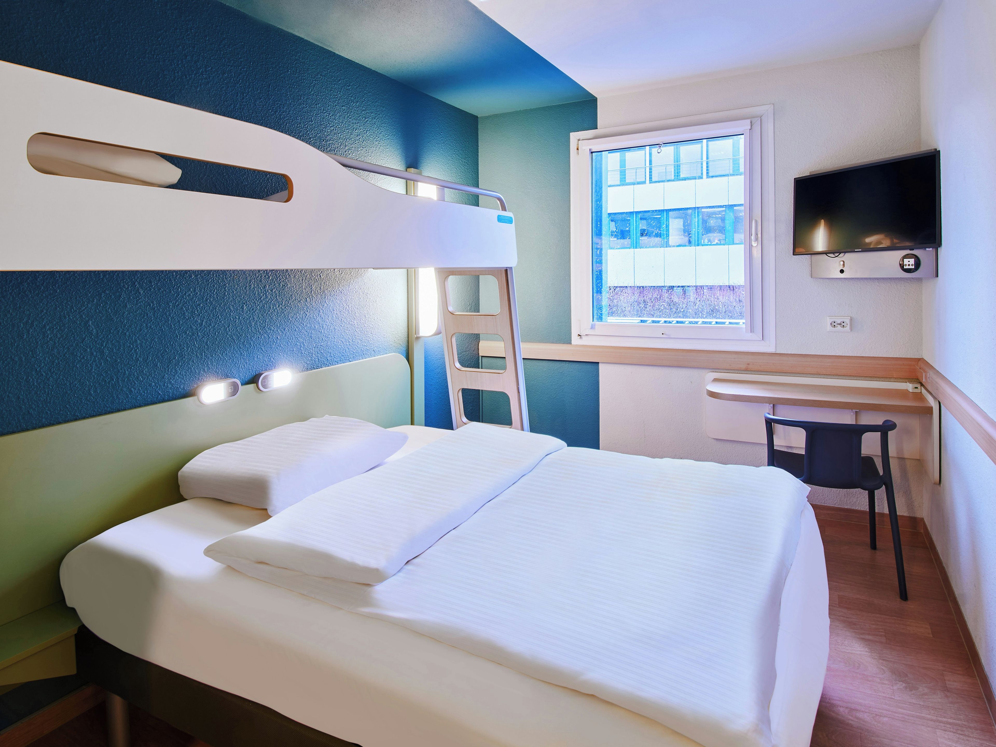 A bunk bed with a ladder, a desk with a chair, a TV mounted on the wall, and a window with a view of buildings.