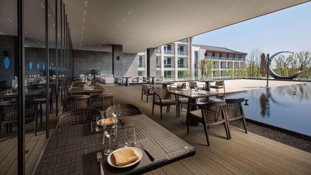 Zona pranzo all'aperto con tavoli e sedie, una piscina e un edificio sullo sfondo.