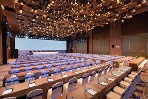 Un salón de conferencias con filas de mesas y sillas, una pantalla grande y un escenario.