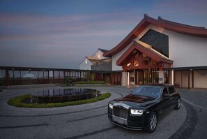 Un bâtiment moderne doté d’un toit incurvé et de grandes fenêtres, une voiture de luxe noire garée devant, et une fontaine circulaire.