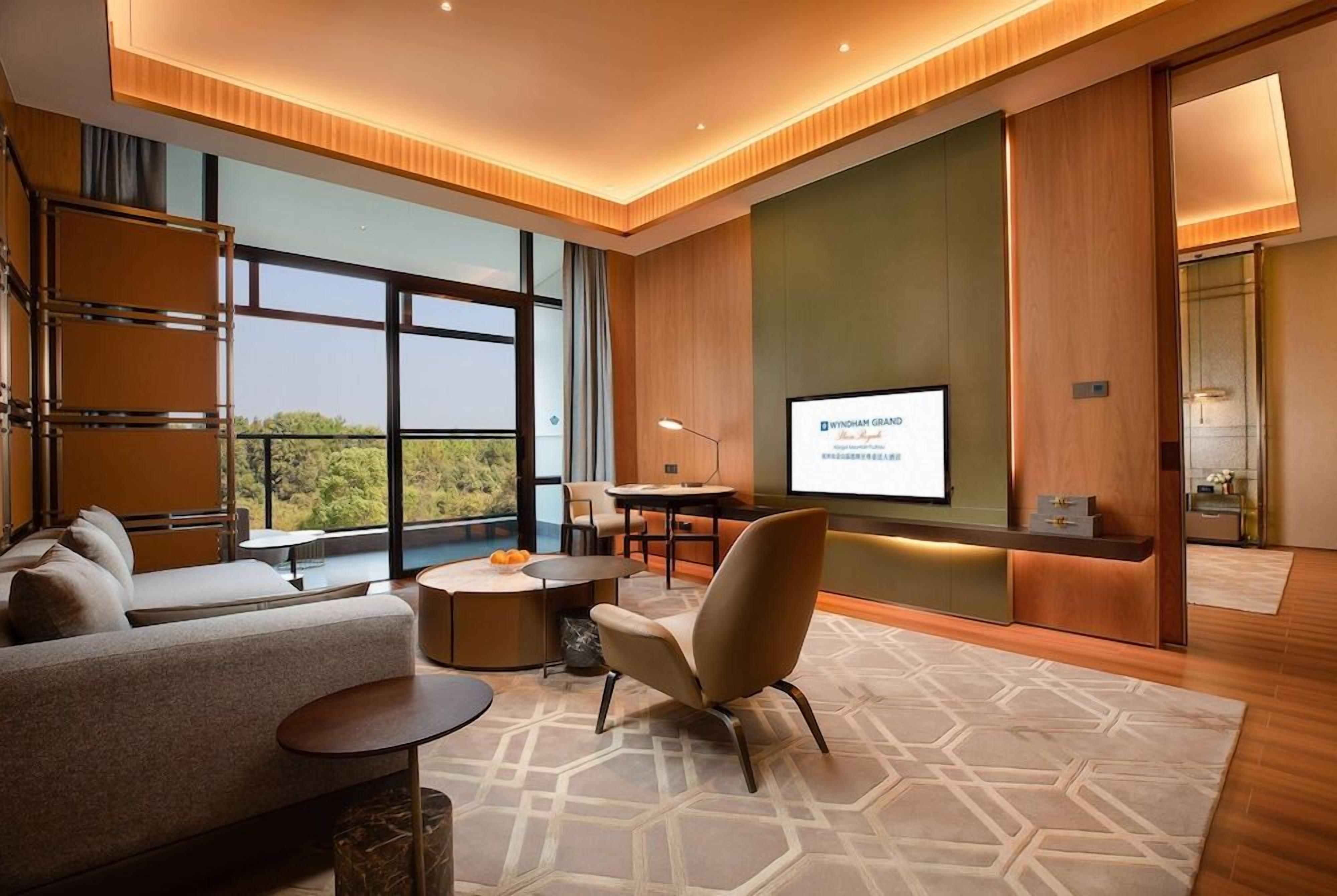 A modern hotel room with a large window, a sofa, a chair, a small table, and a TV screen displaying a message.