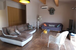 Een woonkamer met een hoekbank, een kleinere bank, een transparante salontafel, een rieten fauteuil en een televisie.