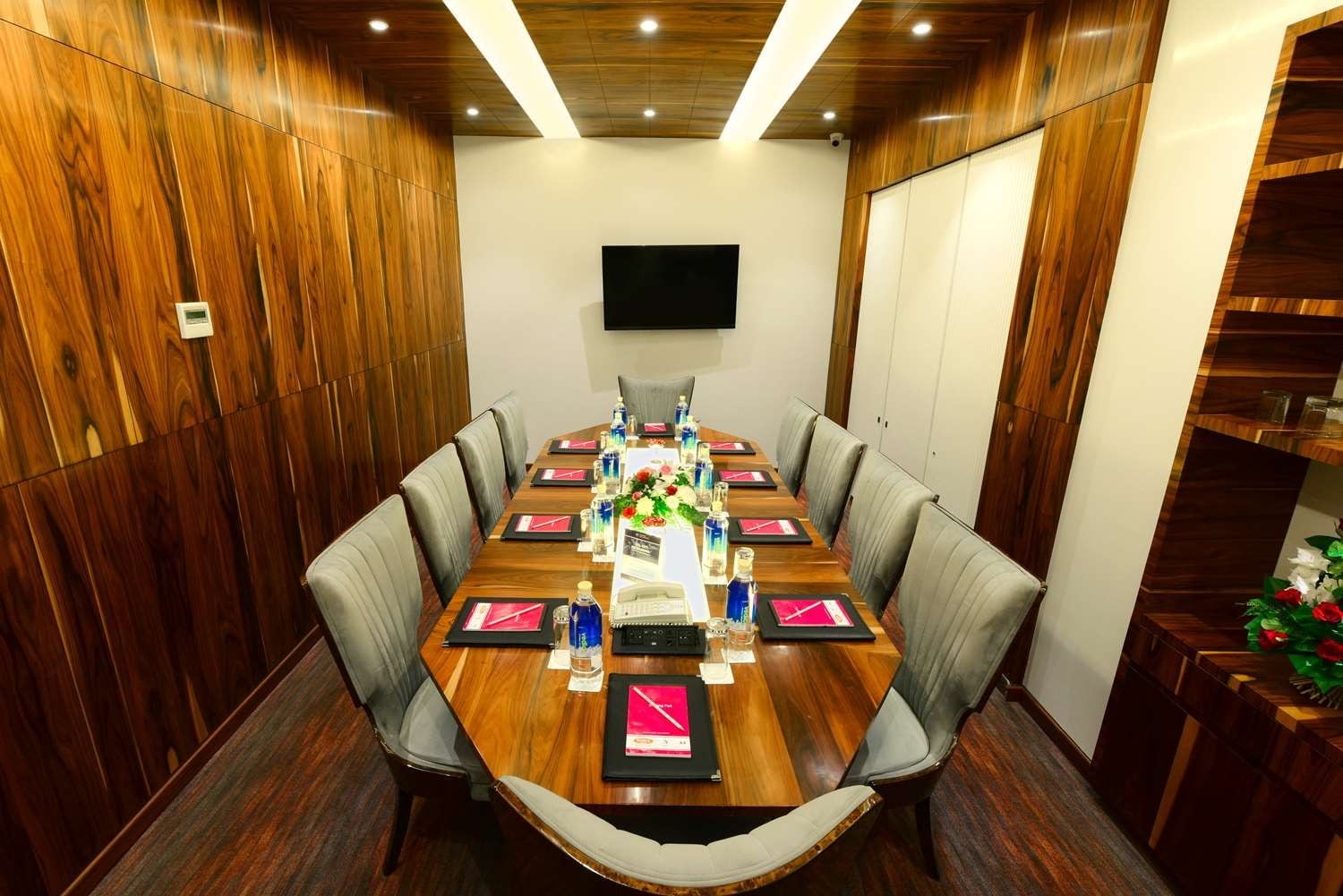 Un salón de conferencias con una larga mesa de madera, sillas tapizadas en gris, un televisor de pantalla plana en la pared y una planta decorativa a la derecha.
